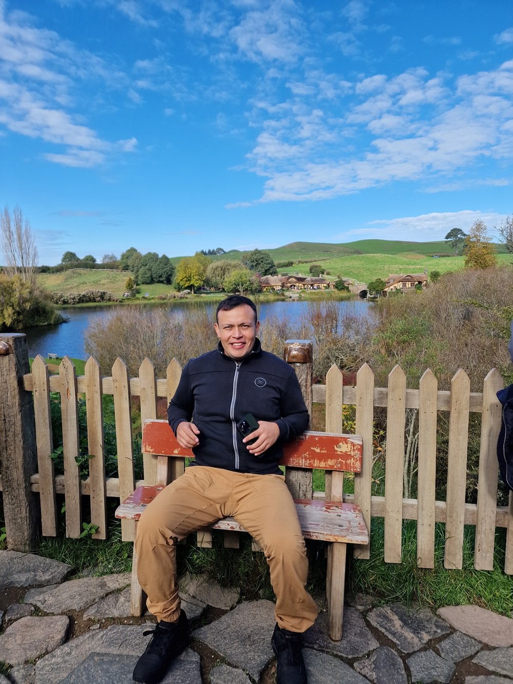 Edgar sentado en una banca frente al lago de Hobbiton, en Nueva Zelanda